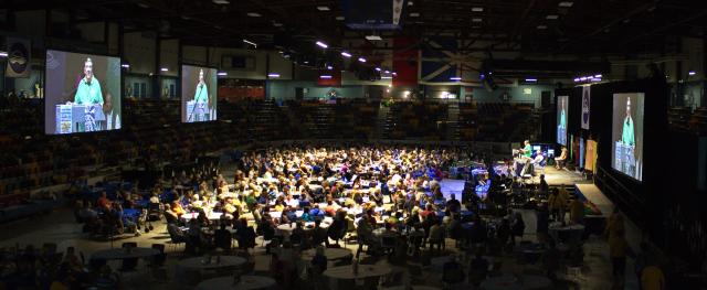 Wide shot of council floor with tables filled with participants, stage with speaker, and screens