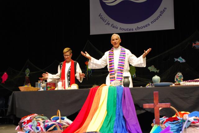 Moderators with open hands speaking in front of a communion table with rainbow flag in front